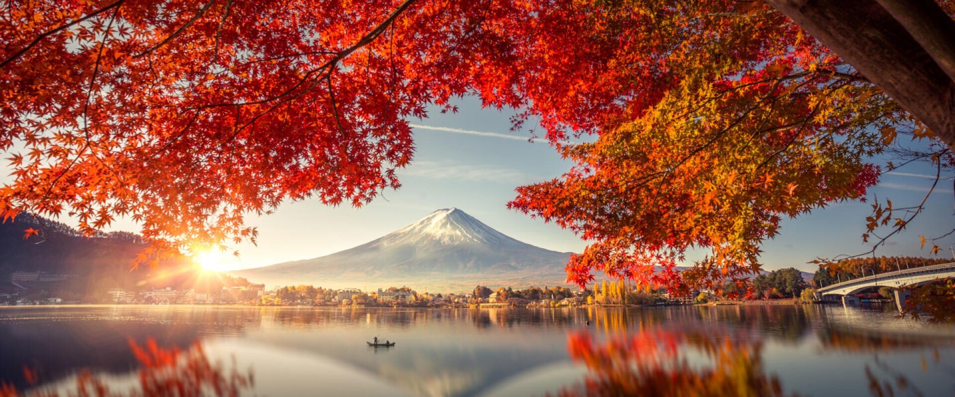 Im Hintergrund erhebt sich der Berg Fuji, umrahmt von leuchtend roten und orangefarbenen Herbstblättern, in denen sich ein ruhiger See spiegelt, in dessen Mitte ein kleines Boot schwimmt. Die Sonne steht tief am Horizont und wirft ein warmes Licht.