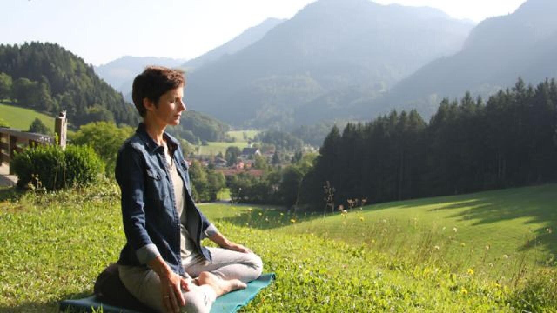Eine Frau sitzt im Schneidersitz auf einer Matte und meditiert im Freien auf einem grasbewachsenen Hügel mit Bergen, Bäumen und einem Tal im Hintergrund unter einem klaren Himmel.